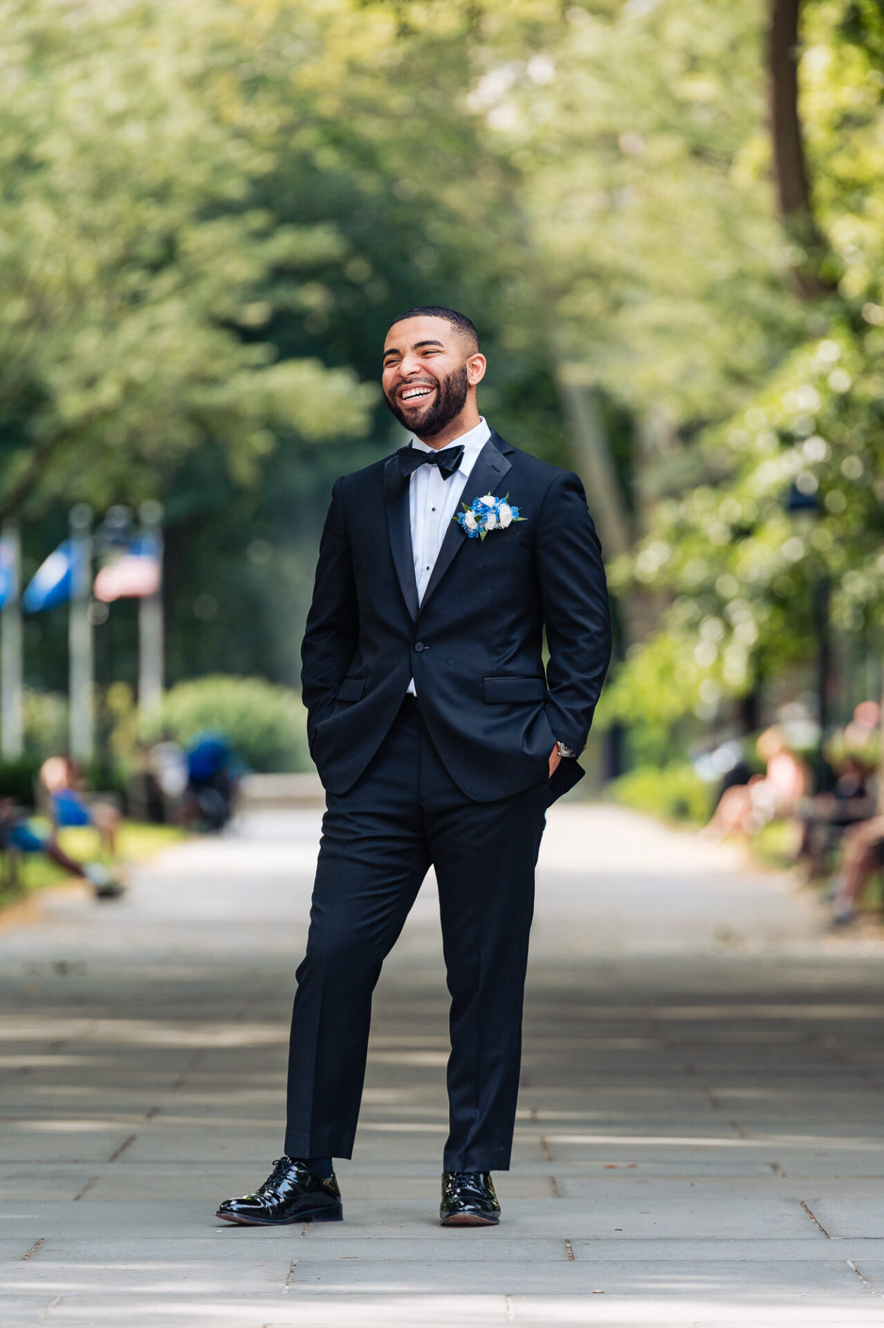 Groom smiling outdoors in a black tuxedo with a black bow tie, blue floral boutonniere, and polished dress shoes on a tree-lined walkway.