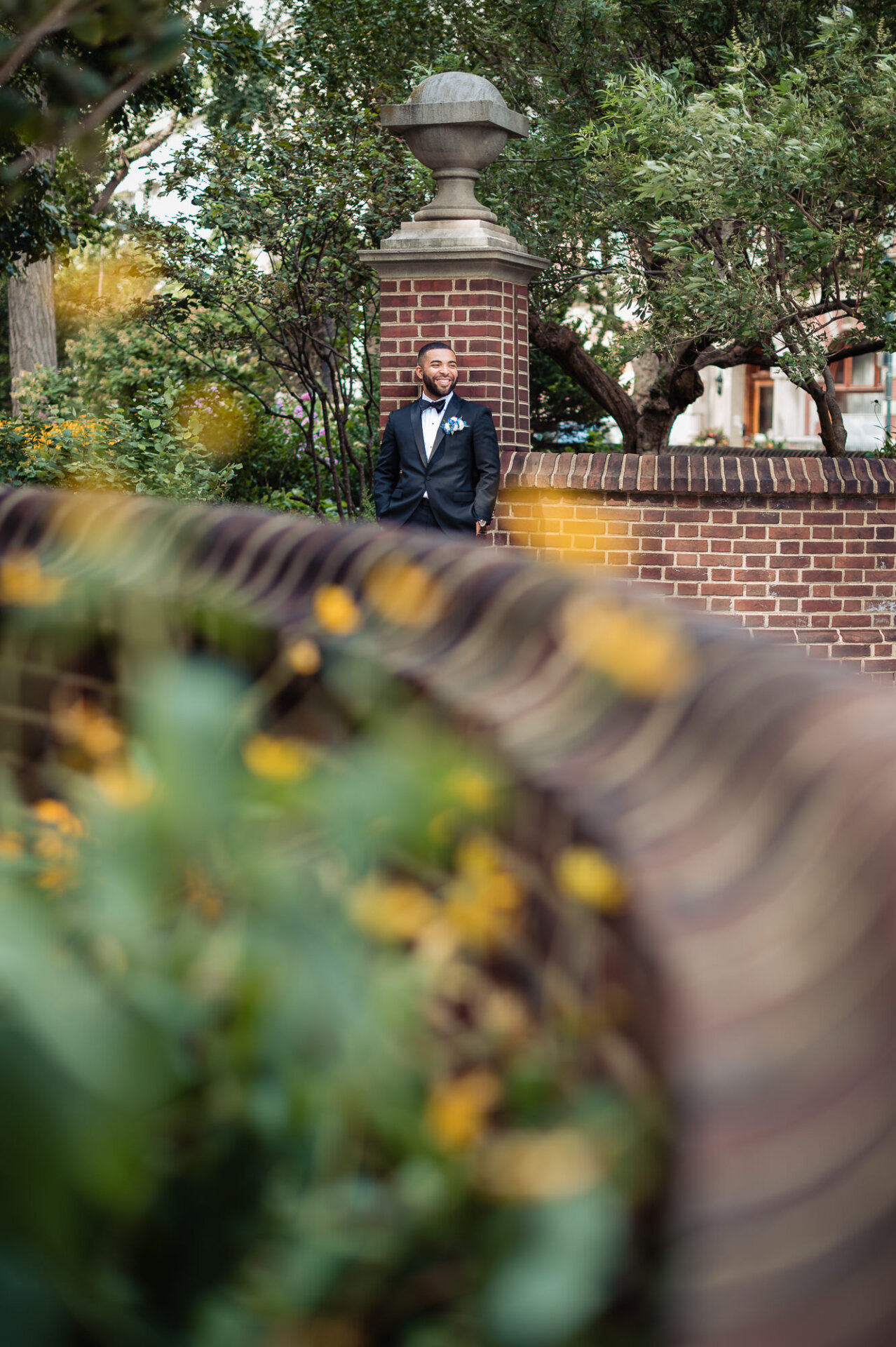 Groom in a black tuxedo and bow tie smiling leaning against a brick wall in a garden setting, framed by blurred yellow flowers in the foreground.