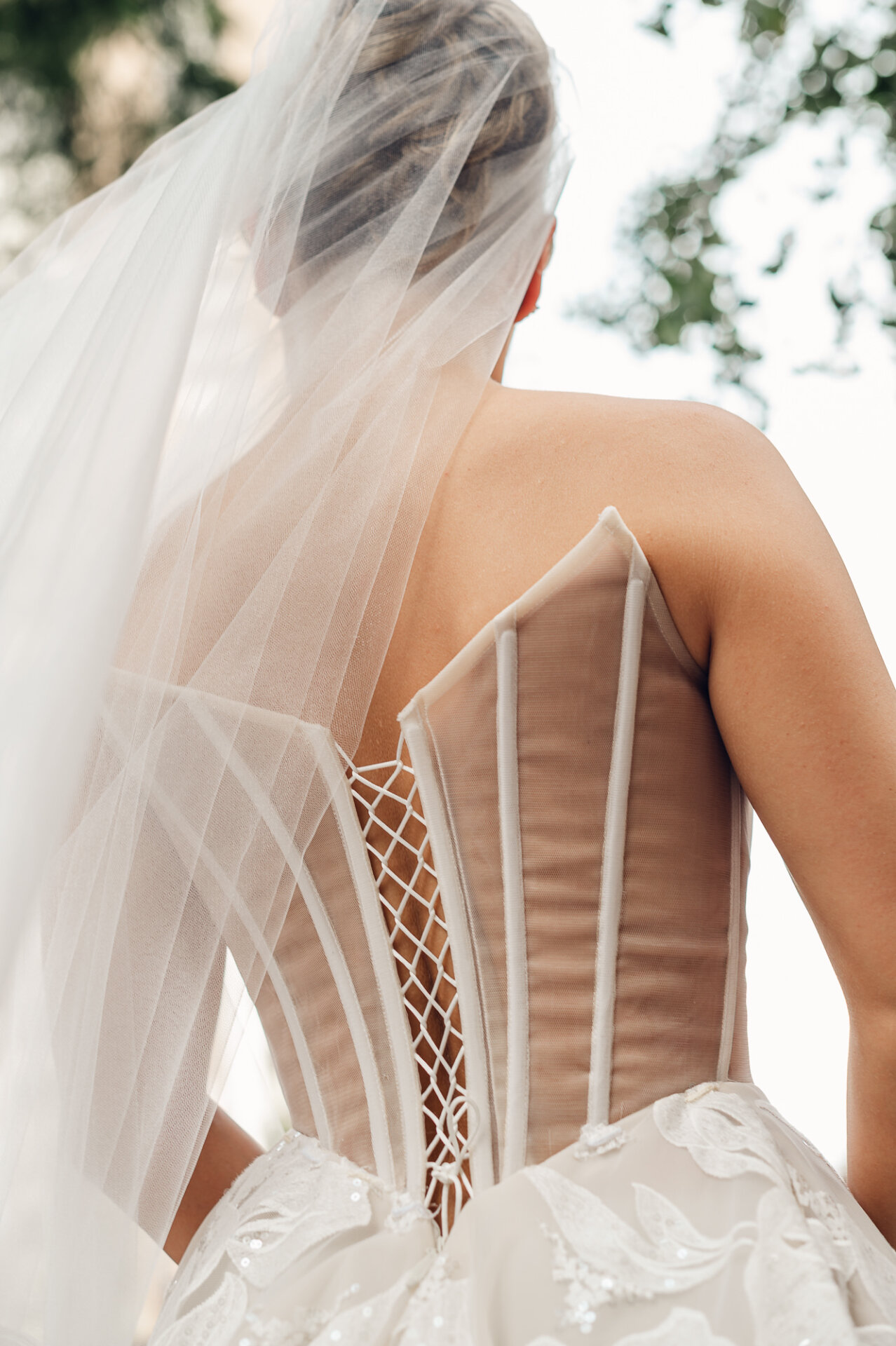Close-up of the back of a bride's wedding dress, showcasing a sheer corset bodice with white boning, lace-up closure, floral embroidery, and a sheer flowing veil.