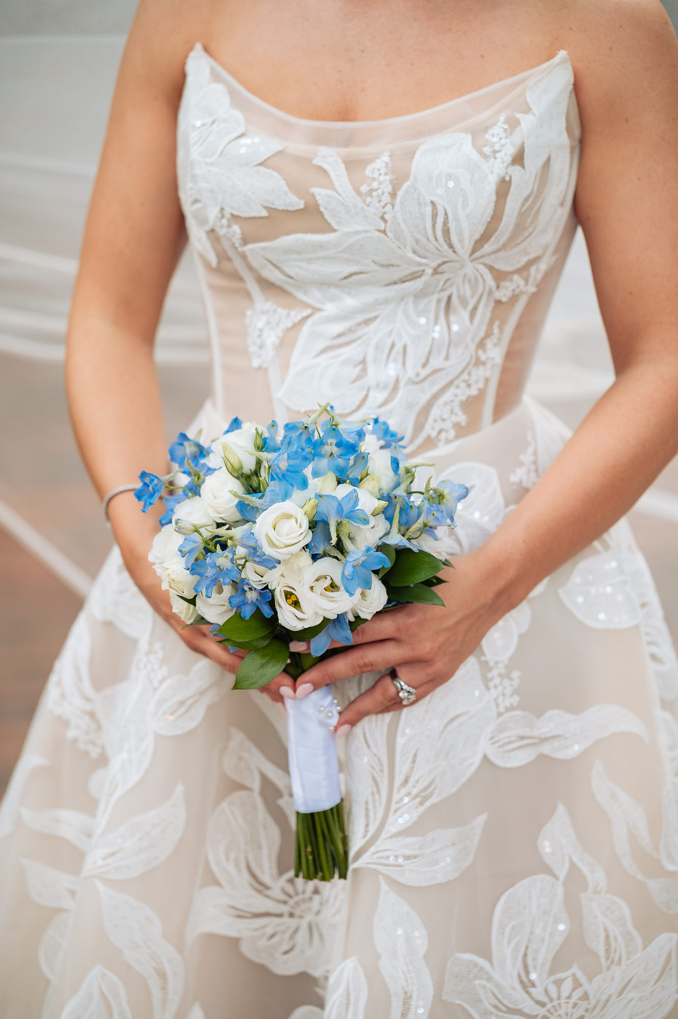 Close-up of a bride holding a round bridal bouquet featuring bright blue delphinium and white roses wrapped in a white satin ribbon.