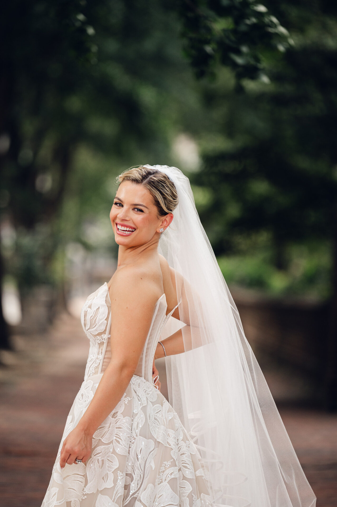 Smiling bride poses outdoors in a floral lace wedding gown and long veil.