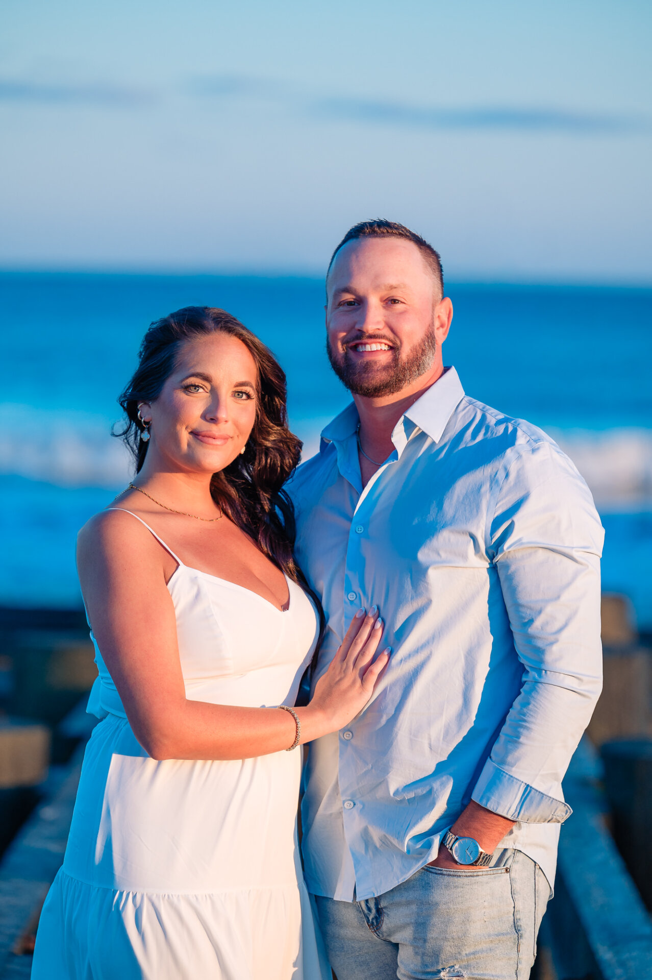 Couple smiling at the camera during their Margate Beach engagement session, with blue ocean waves in the background