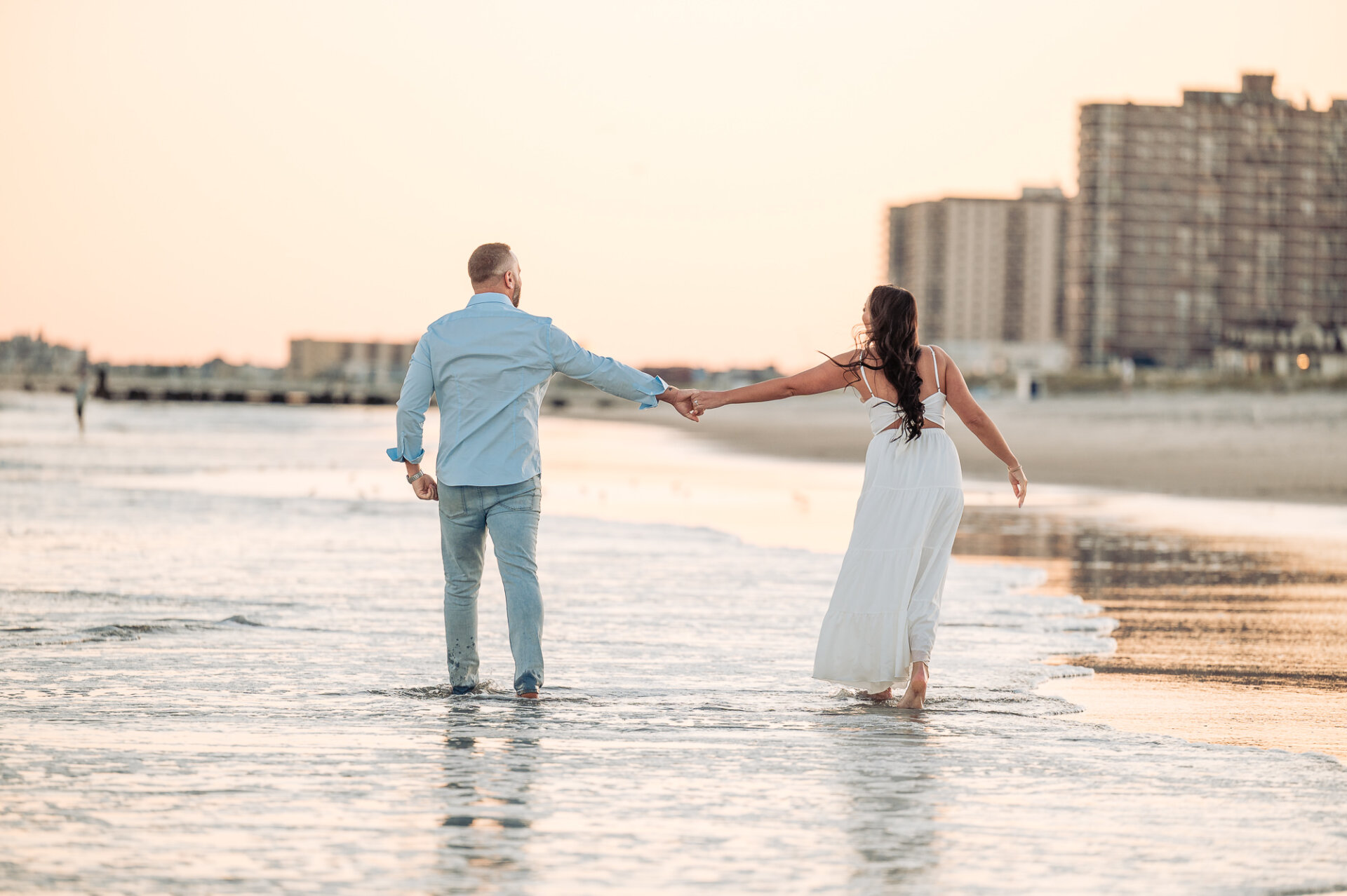 Couple holding hands while walking barefoot through shallow ocean water at sunset on Margate Beach