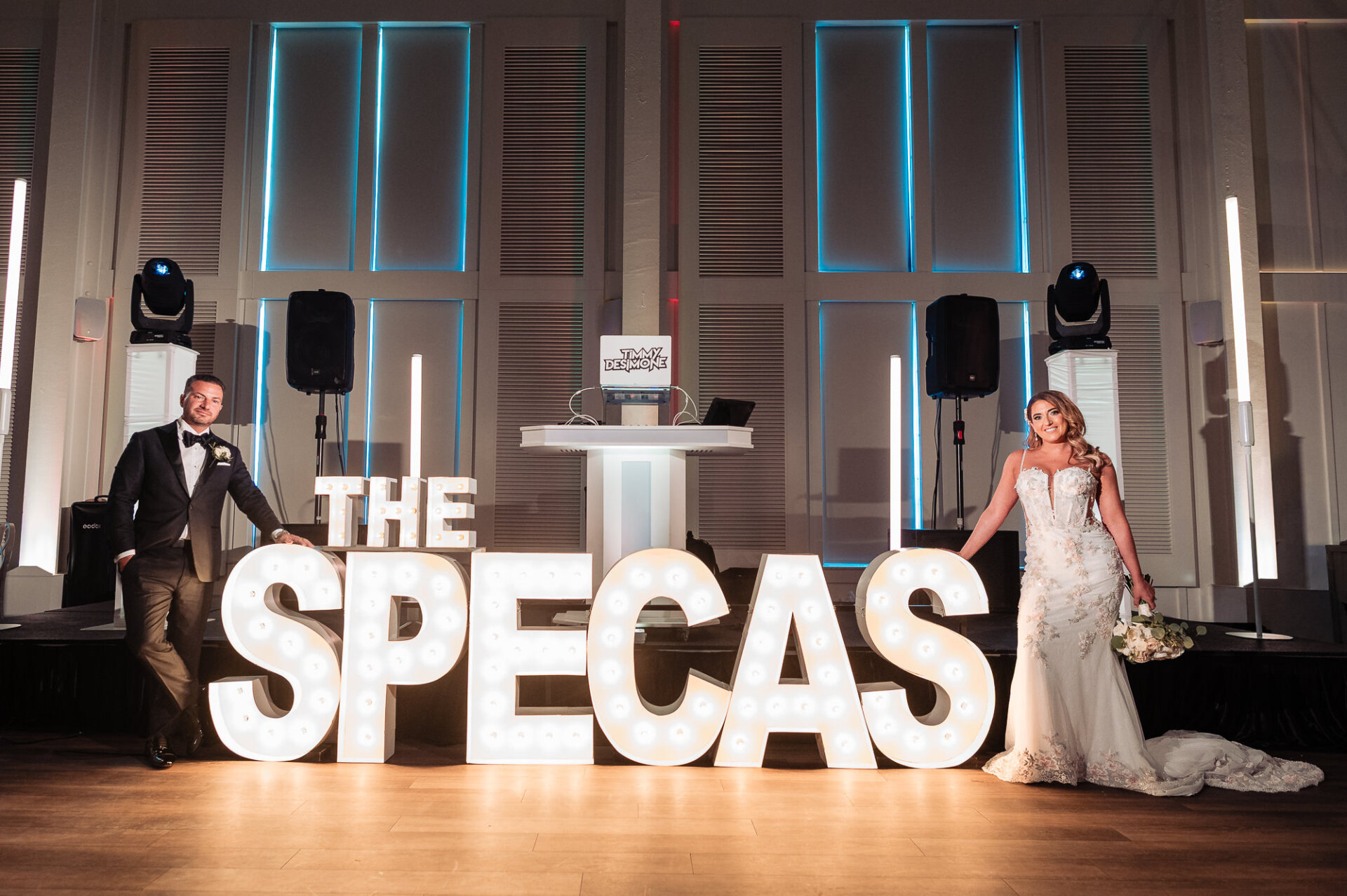 Bride and groom posing on dance floor beside large illuminated marquee letters spelling THE SPECAS