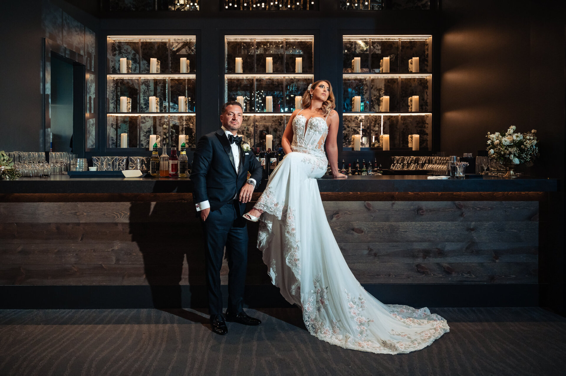 Full length portrait of bride sitting on bar counter and groom standing beside her in front of lit candle shelves