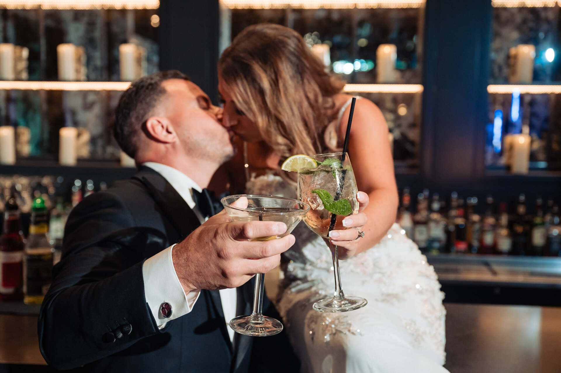 Bride and groom sharing a kiss while holding cocktail drinks at dark reception bar counter