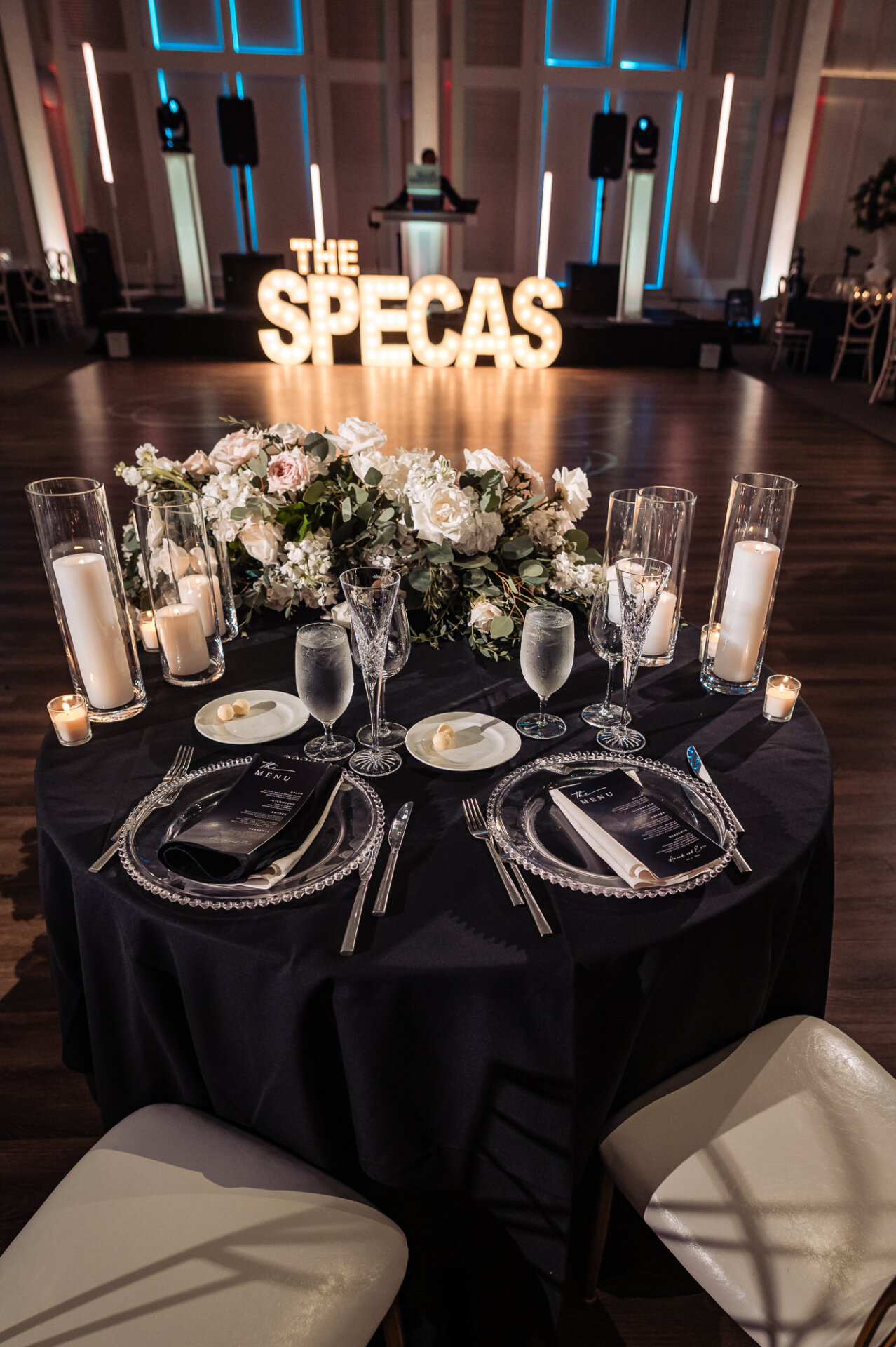 Sweetheart table setup with black tablecloth, white floral arrangement, cylinder candles, and marquee light sign reading THE SPECAS