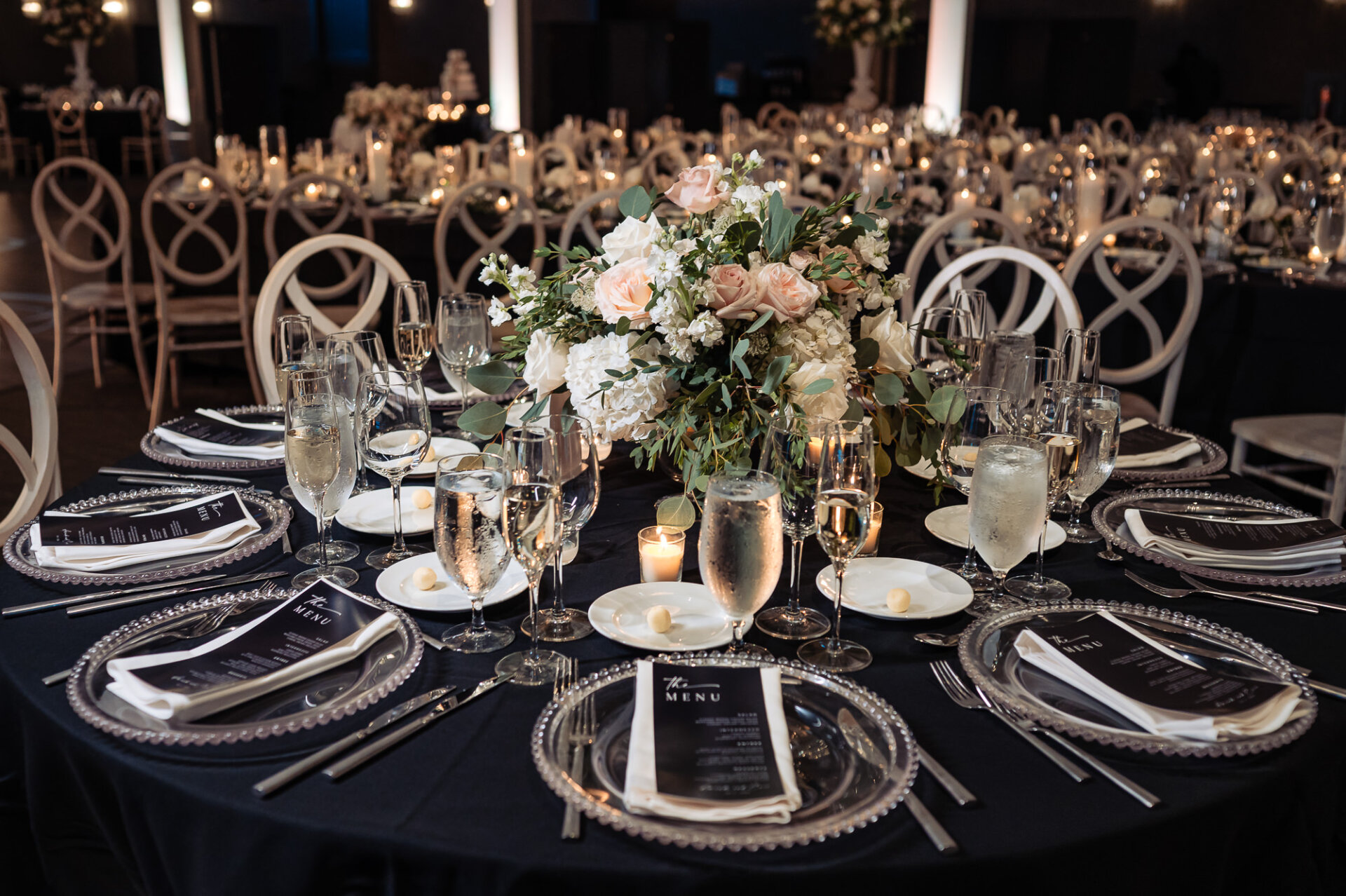 Guest dining table setup with dark linens, white floral centerpiece, and beaded charger plates with black menu cards