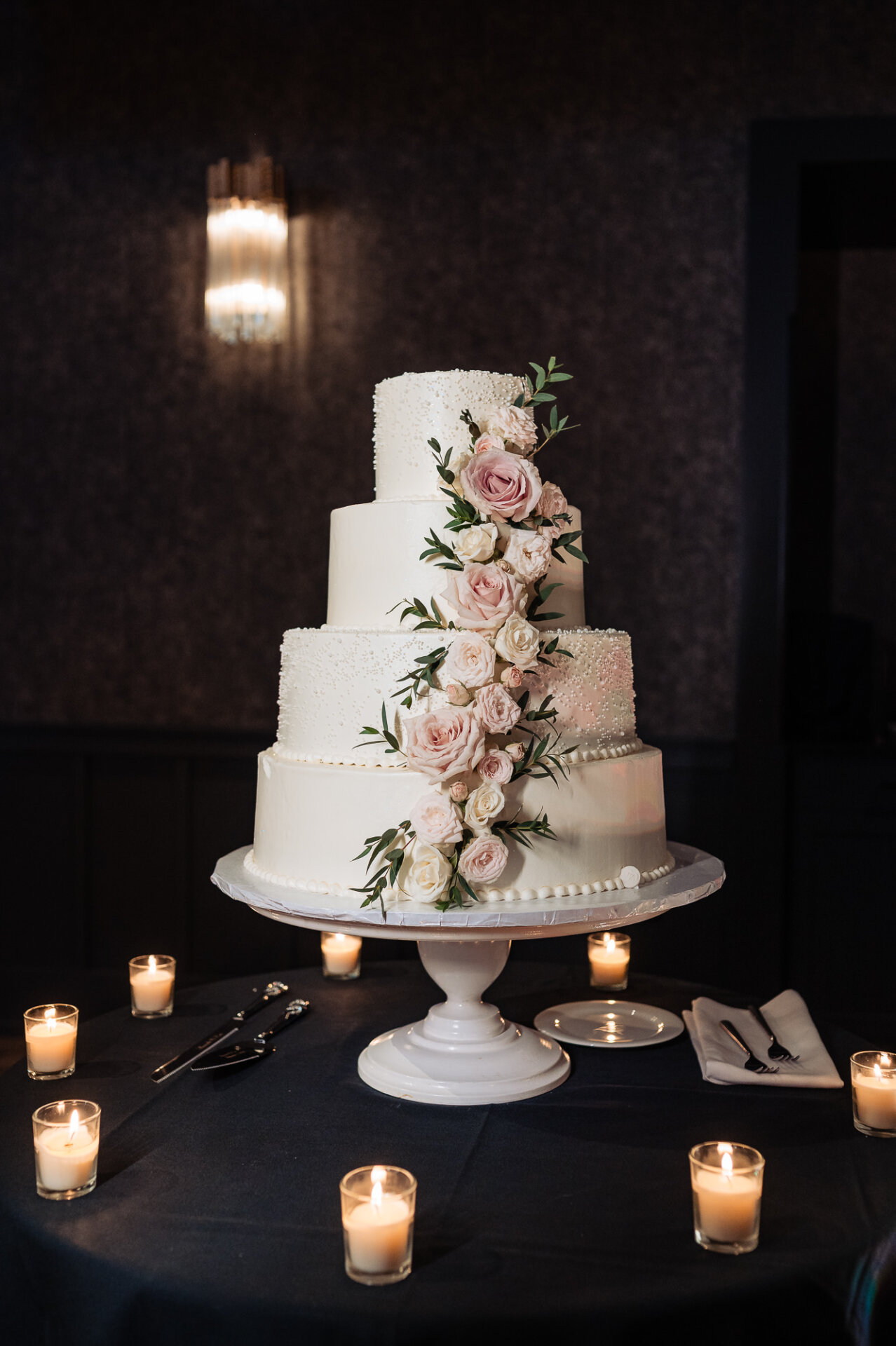 Four-tier white wedding cake decorated with cascading blush pink roses surrounded by votive candles