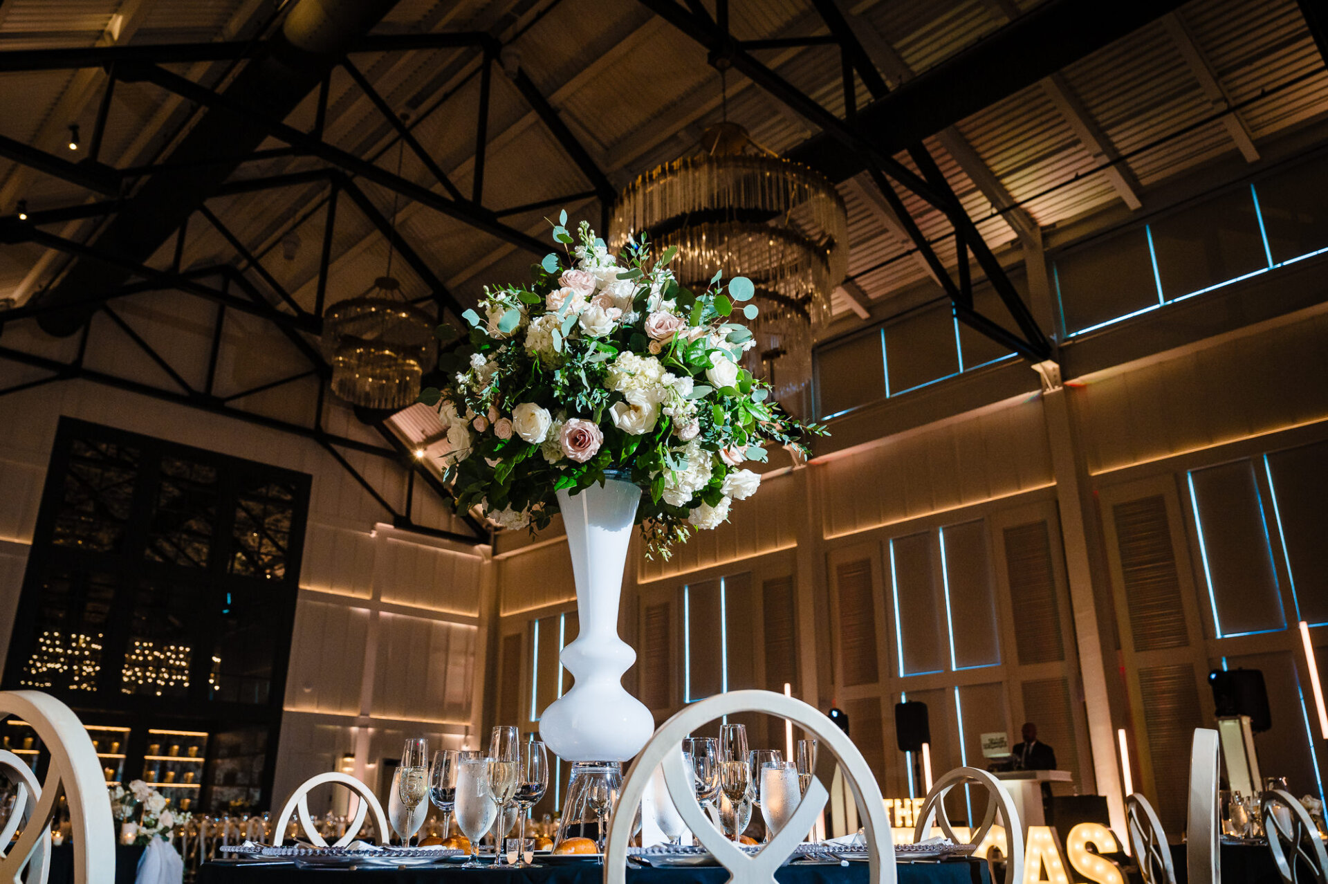 Tall white urn centerpiece with lush floral arrangement on reception dining table under high ceilings