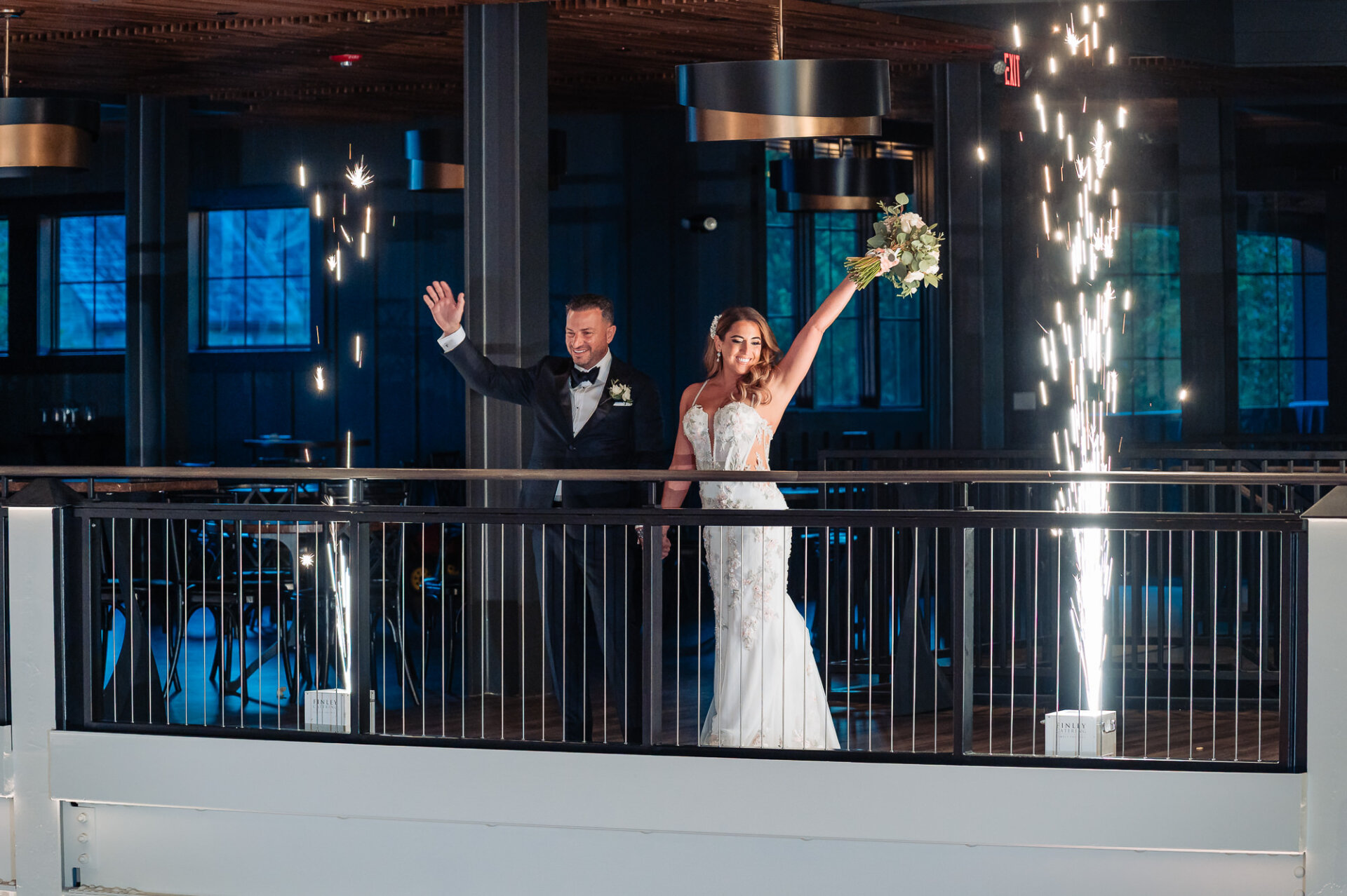 Newlyweds grand entrance on indoor balcony waving to guests while sparkler fountains shoot up