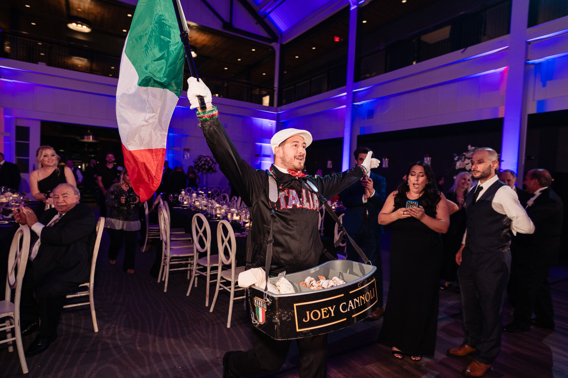 Joey Cannoli vendor in Italian attire holding Italian flag and carrying tray of fresh cannolis through reception