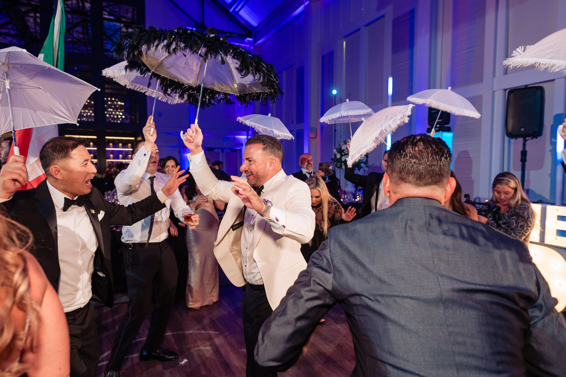 Groom in white tux jacket dancing enthusiastically under black feather parasol with wedding guests