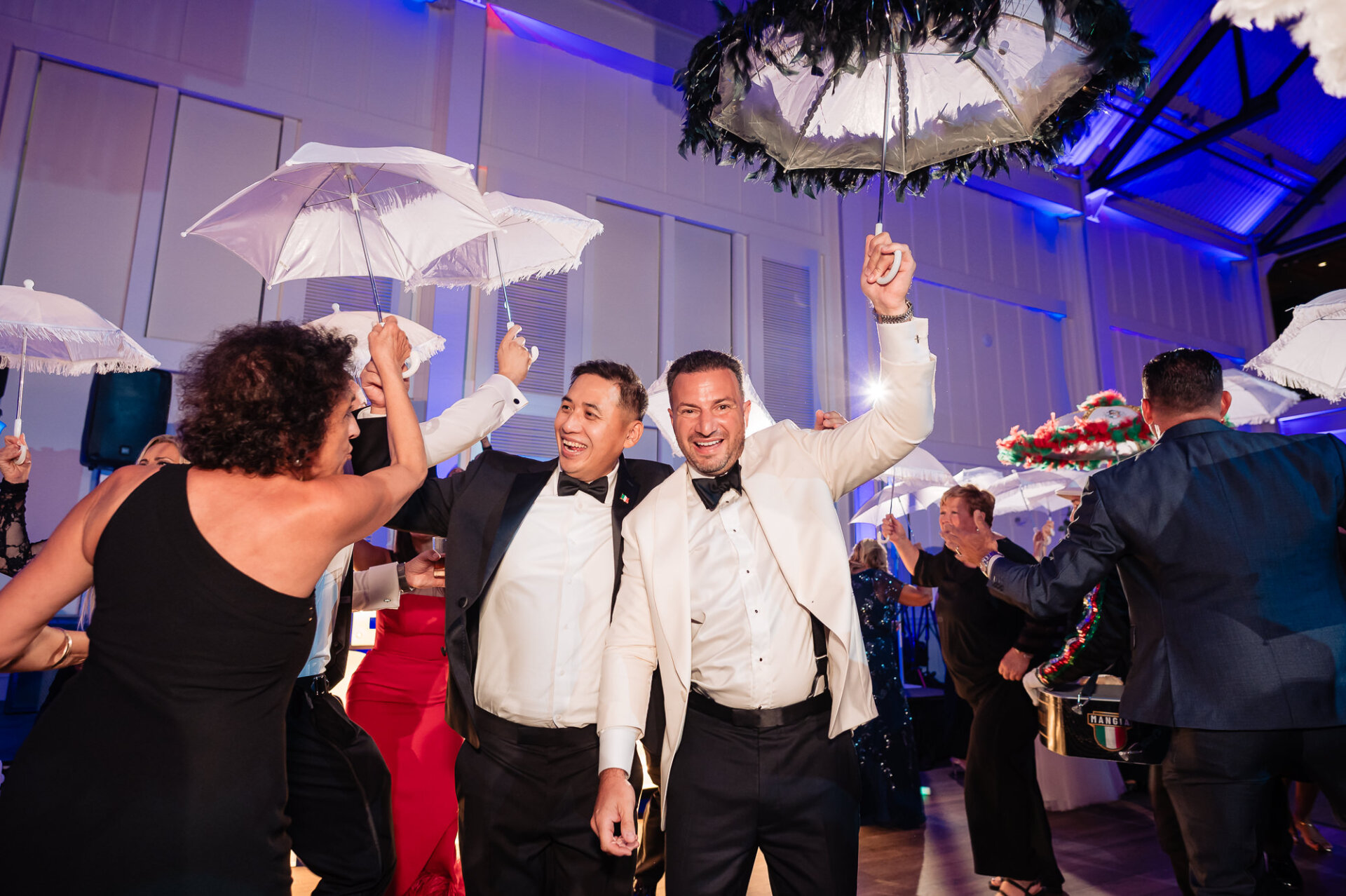 Groom wearing white dinner jacket dancing under feather parasol with smiling wedding guests