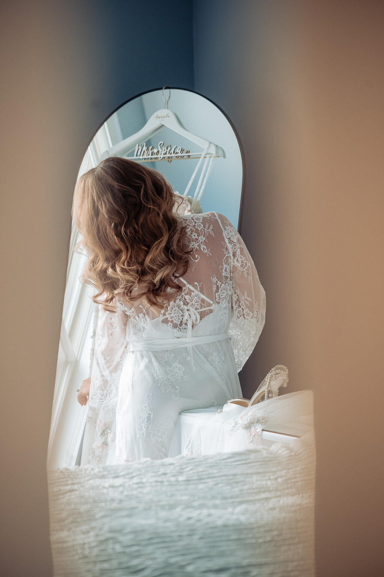 Bride wearing lace robe looking in arched mirror at her wedding gown hanging in background