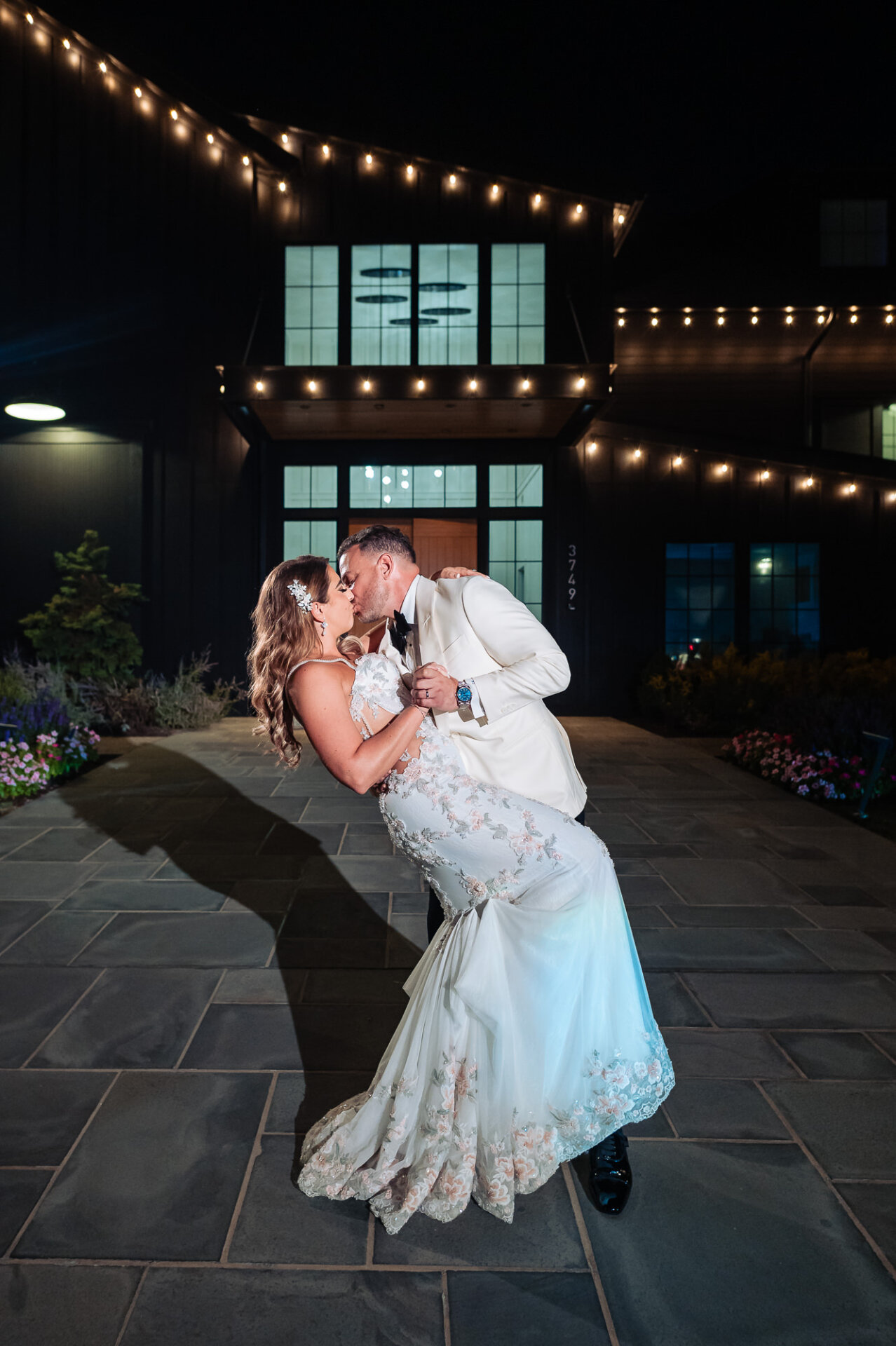 Groom dipping and kissing bride outdoors at night under string lights in front of venue entrance