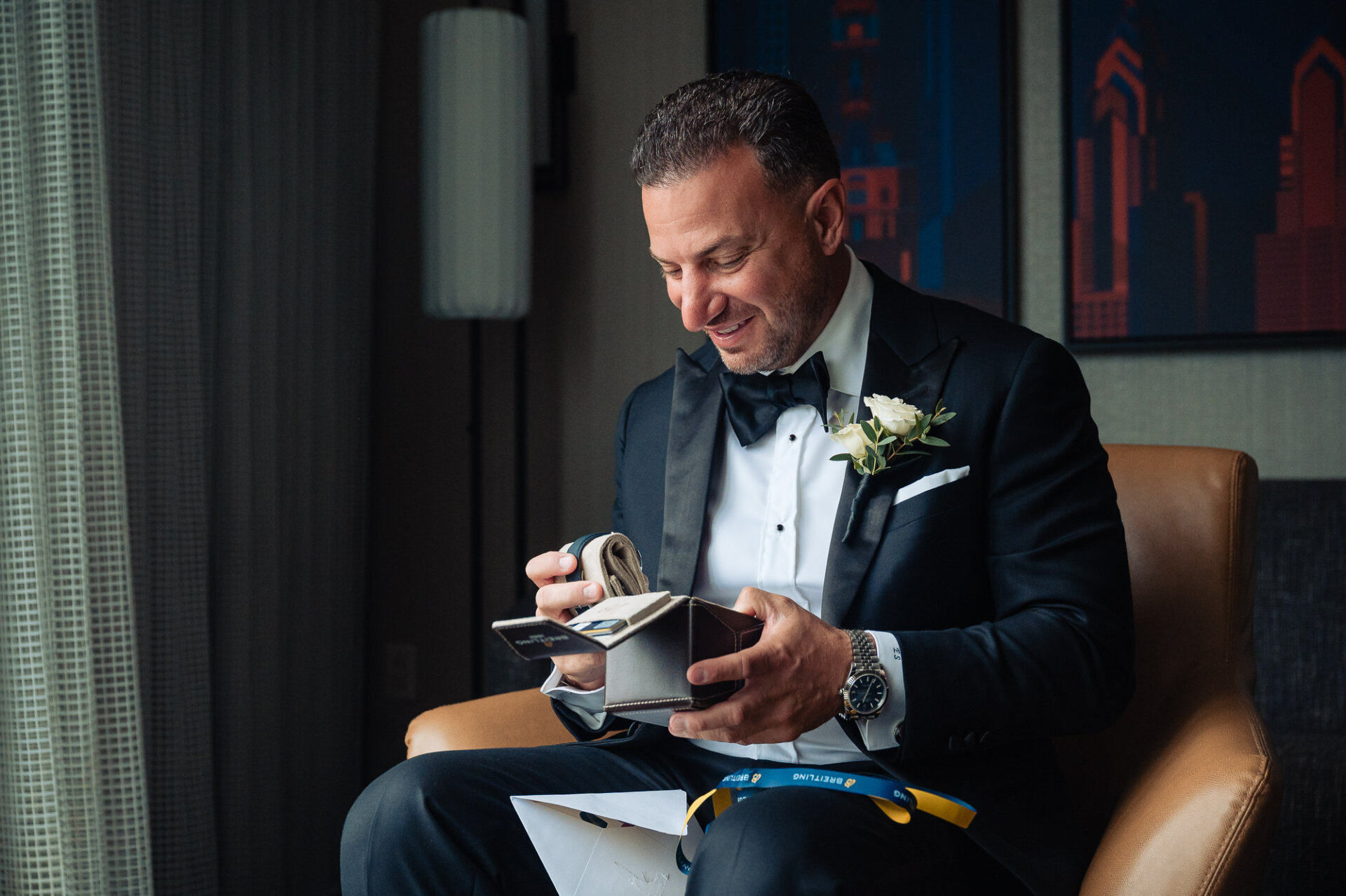 Groom wearing tuxedo unboxing blue Breitling watch gift while sitting in chair