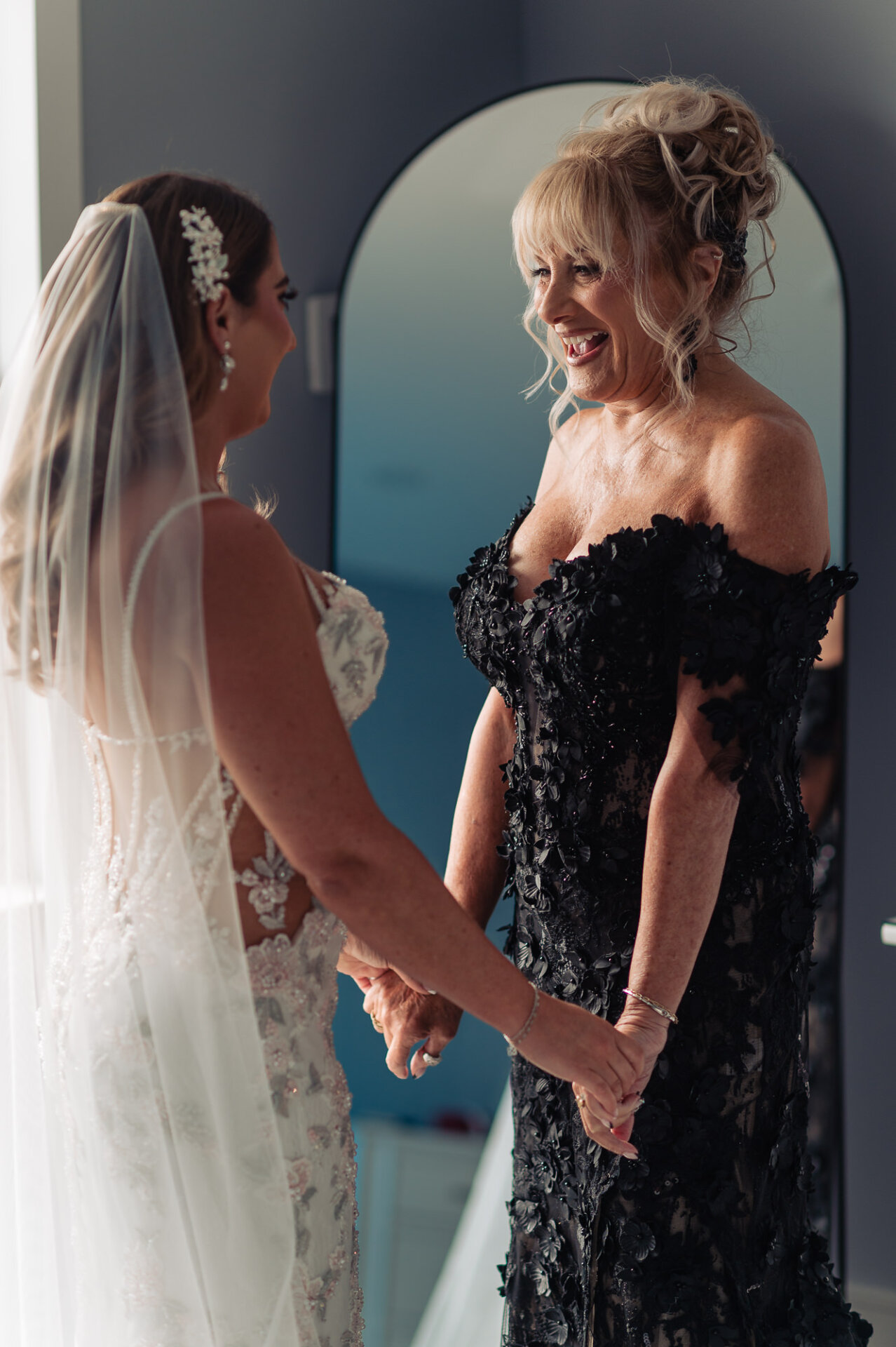 Bride holding hands and laughing with mother of the bride wearing off-the-shoulder black lace gown