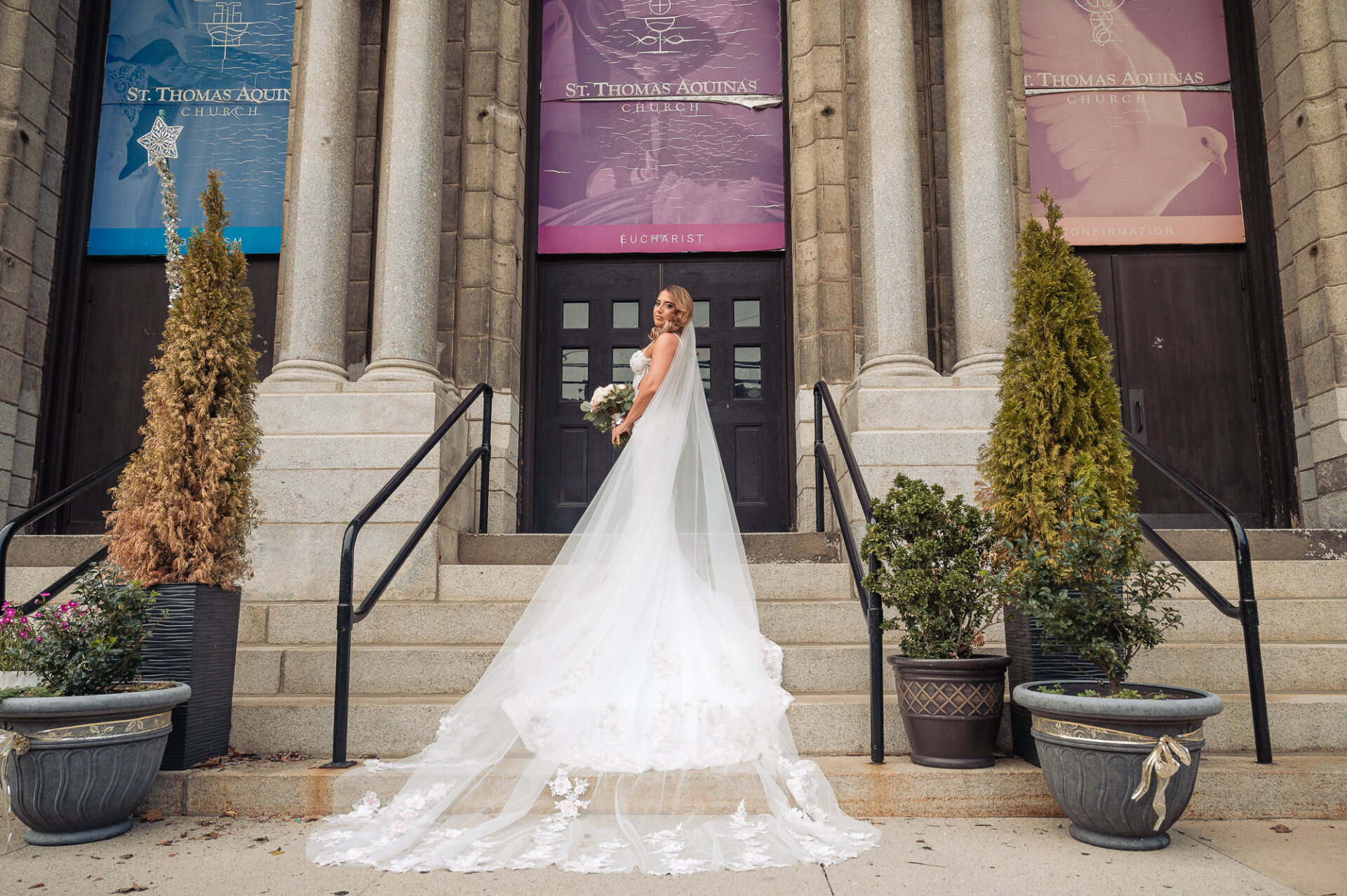Bride with long veil spread down stone steps standing in front of entrance to St. Thomas Aquinas Church
