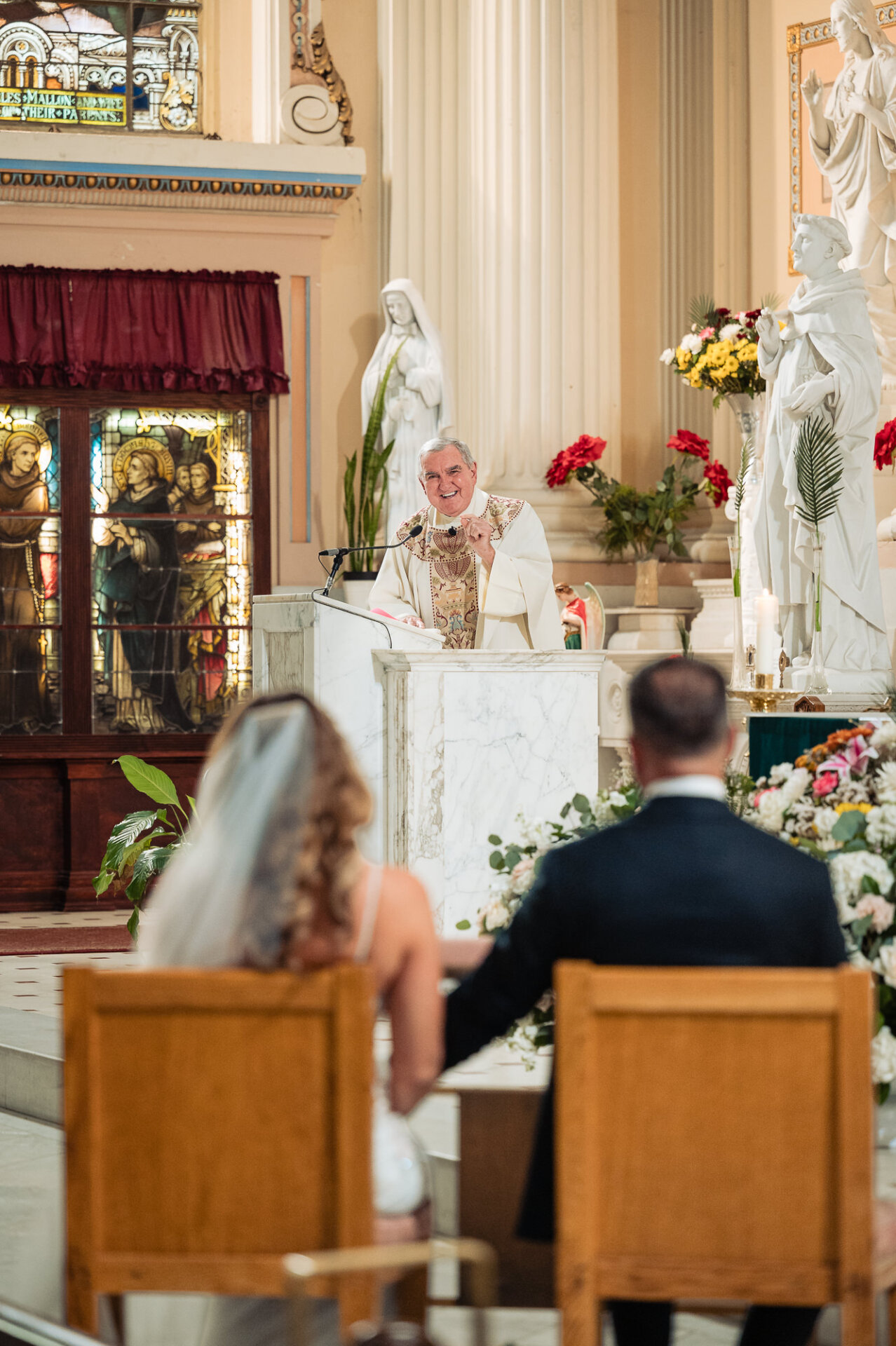 View from behind seated bride and groom listening to priest speak at pulpit during wedding mass
