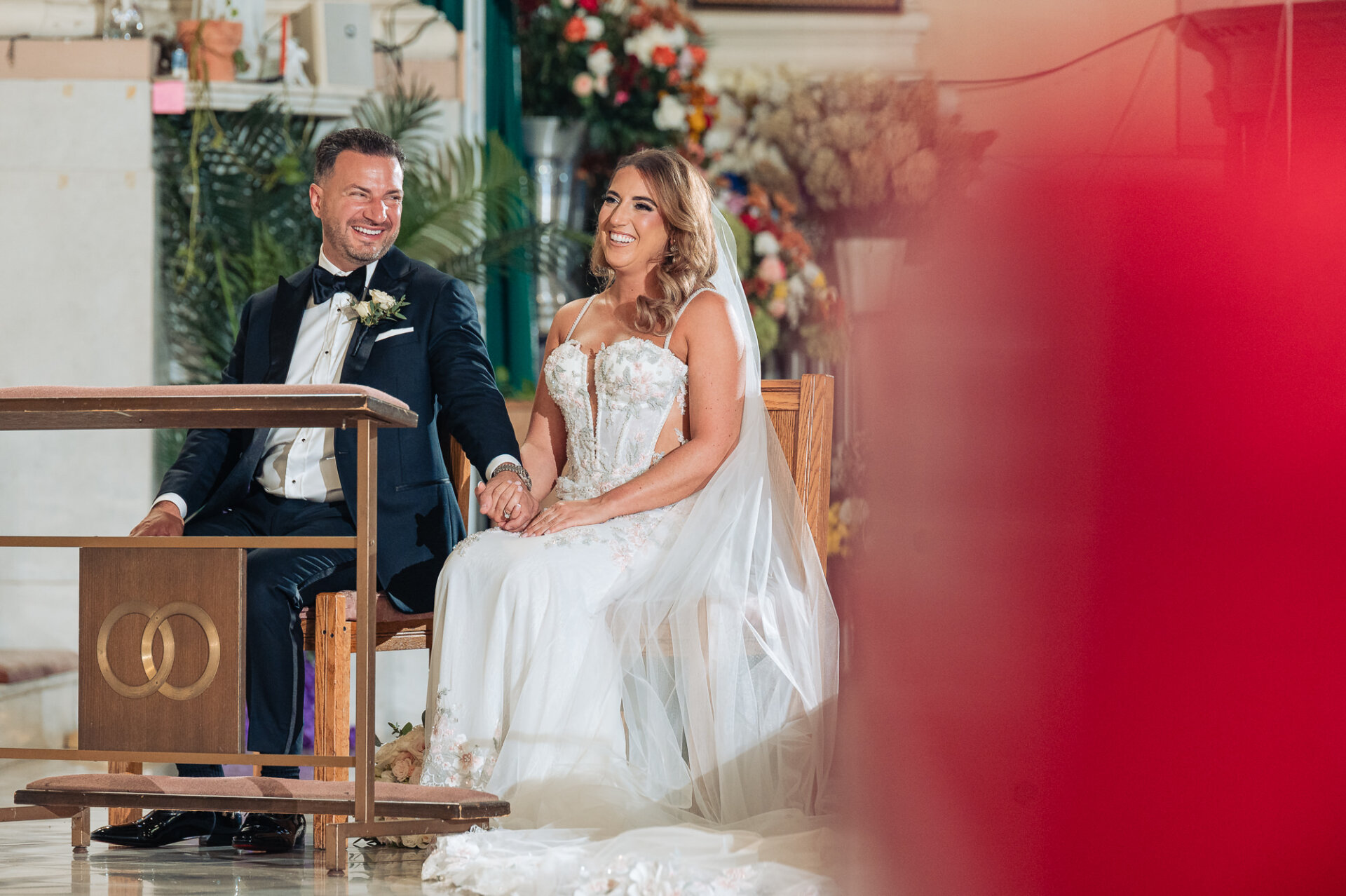 Bride and groom holding hands and smiling while seated at prayer bench during ceremony