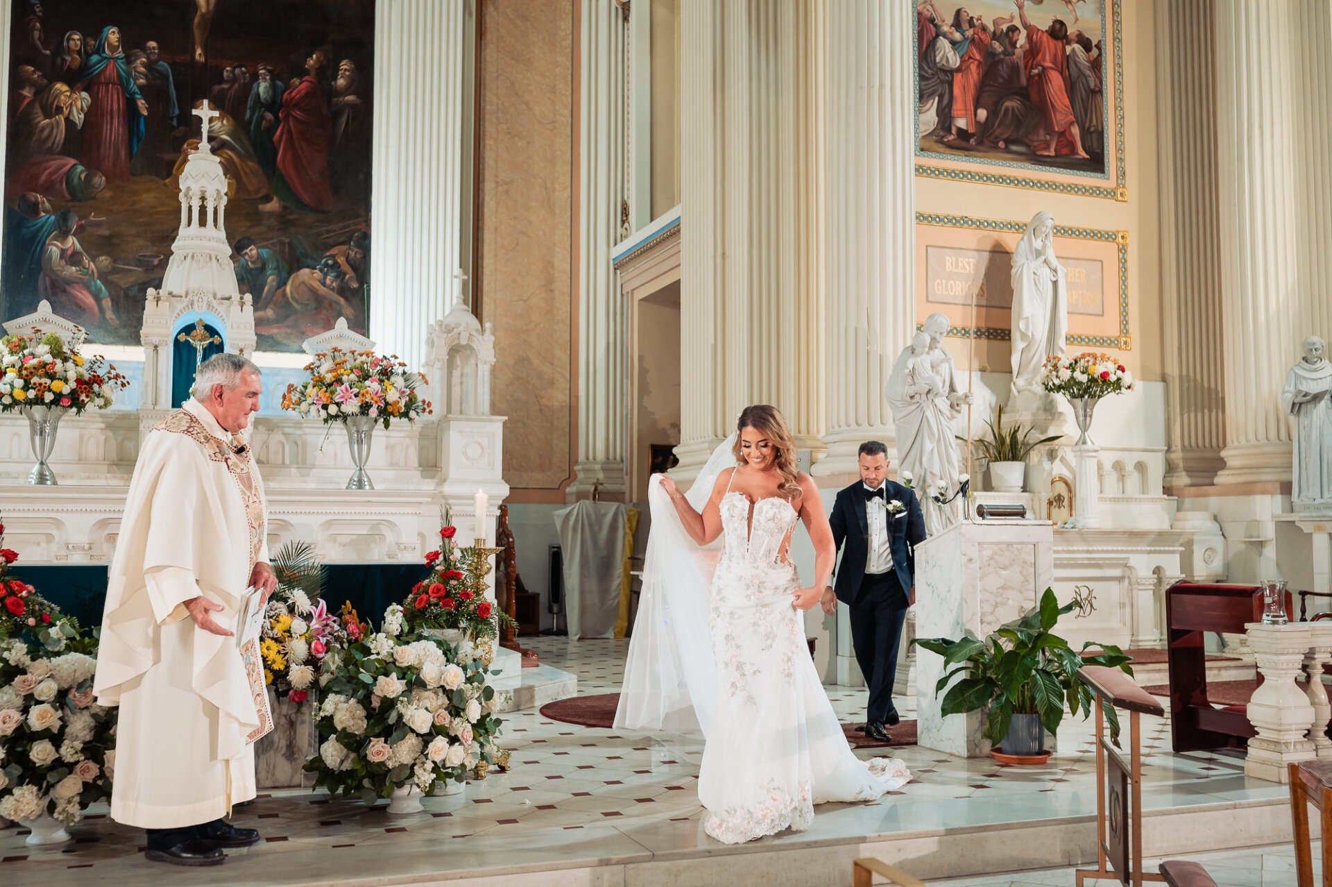 Bride holding train of her wedding dress while walking up altar steps past priest during church ceremony