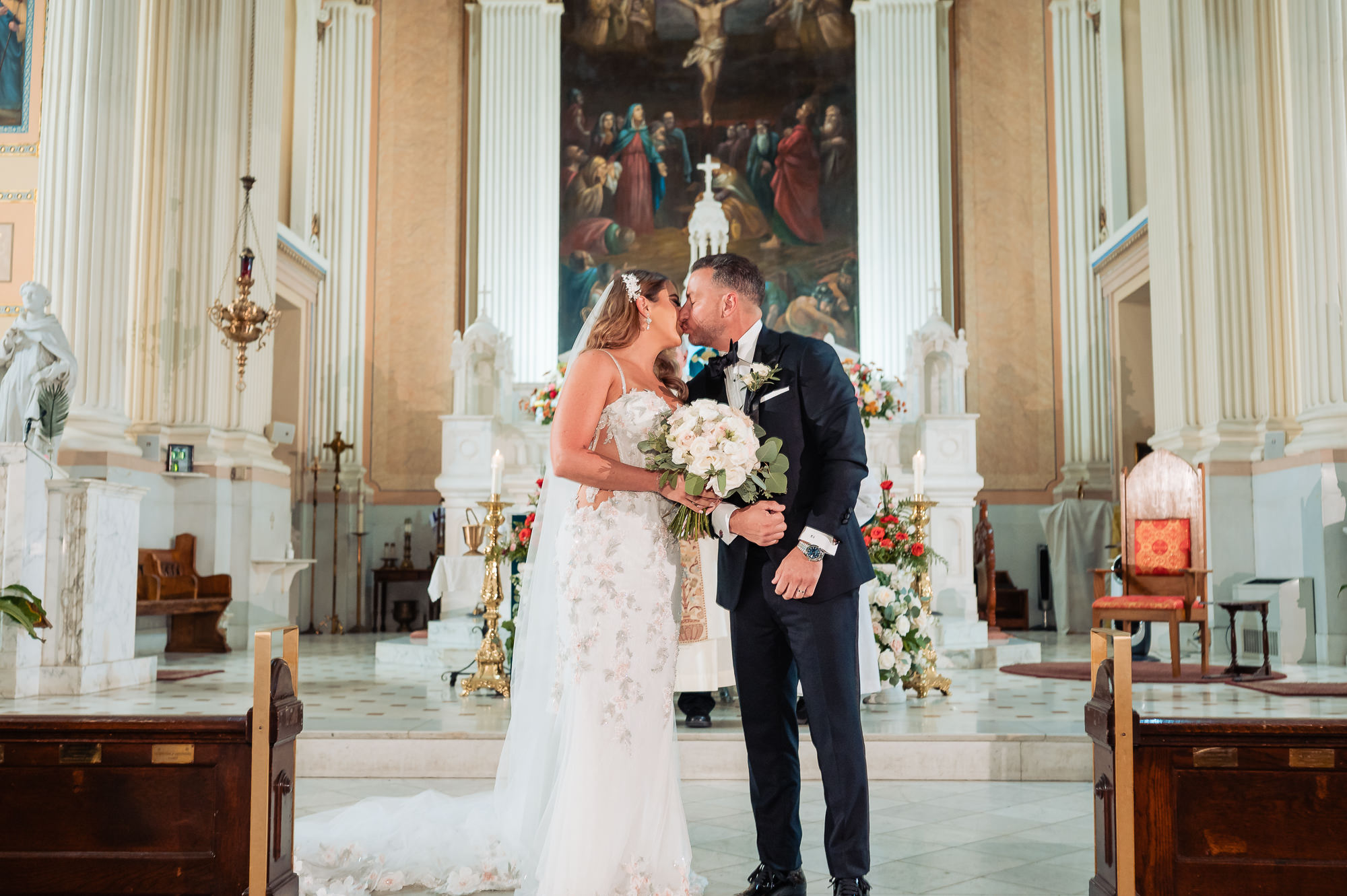 Bride holding white floral bouquet while sharing first kiss with groom at church altar