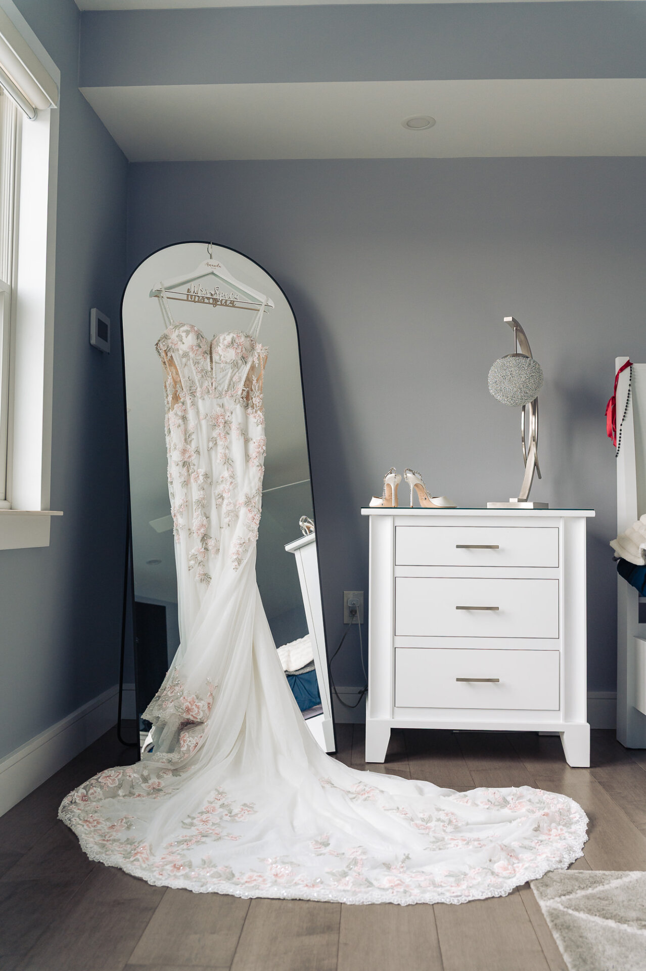 Floral embroidered wedding dress hanging on mirror next to white nightstand with bridal heels