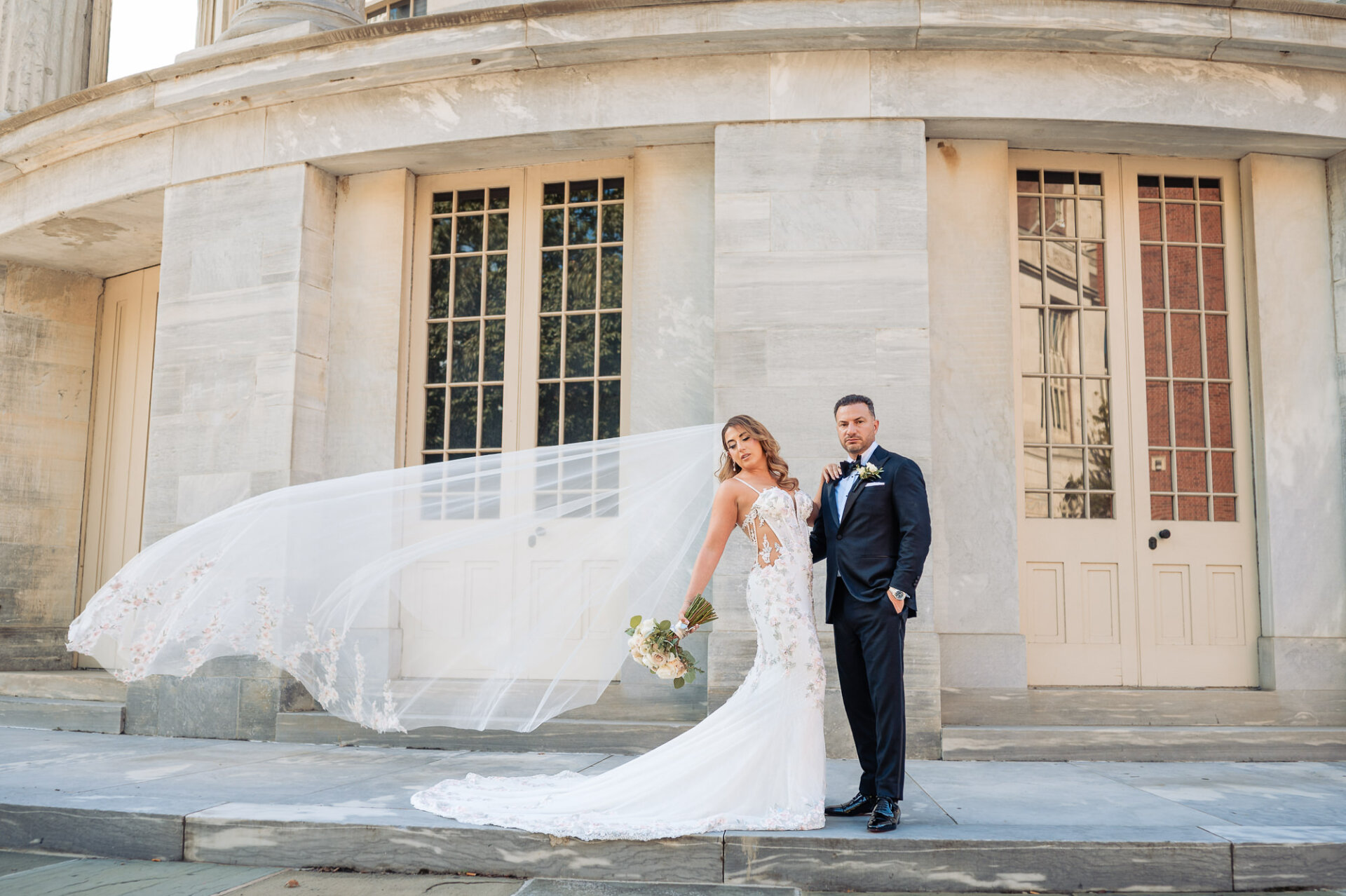 Full length portrait of bride with flowing veil and groom posing in front of stone columns