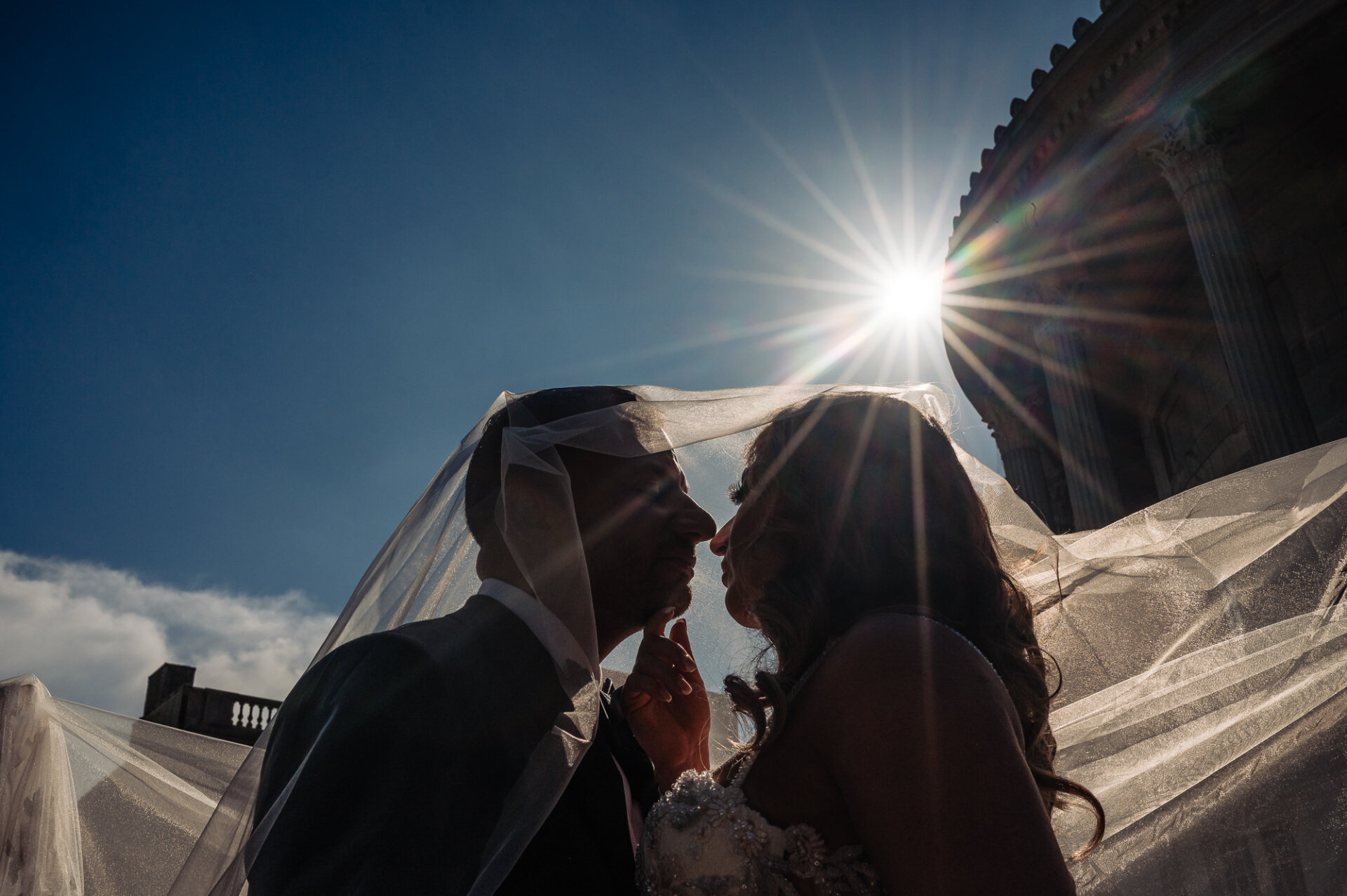 Sunburst silhouette shot of bride and groom embrace under flowing veil