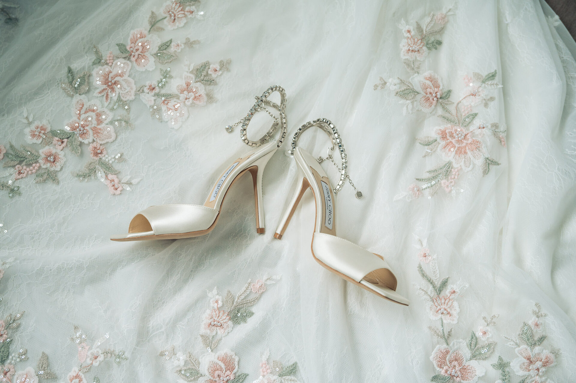 Pair of white satin Jimmy Choo bridal heels with crystal ankle straps on floral lace gown