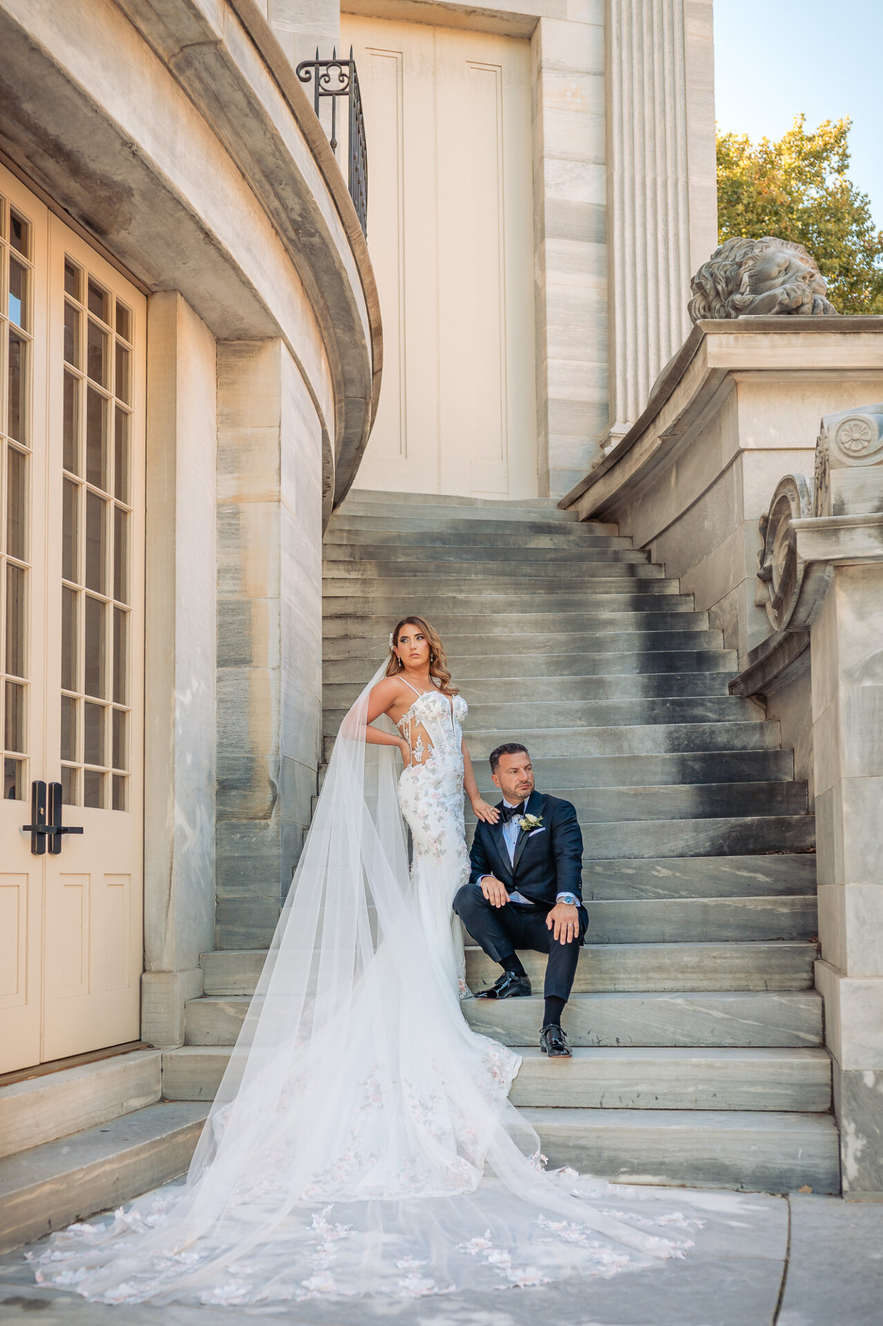 Bride standing and groom sitting on outdoor stone steps in front of historic architecture