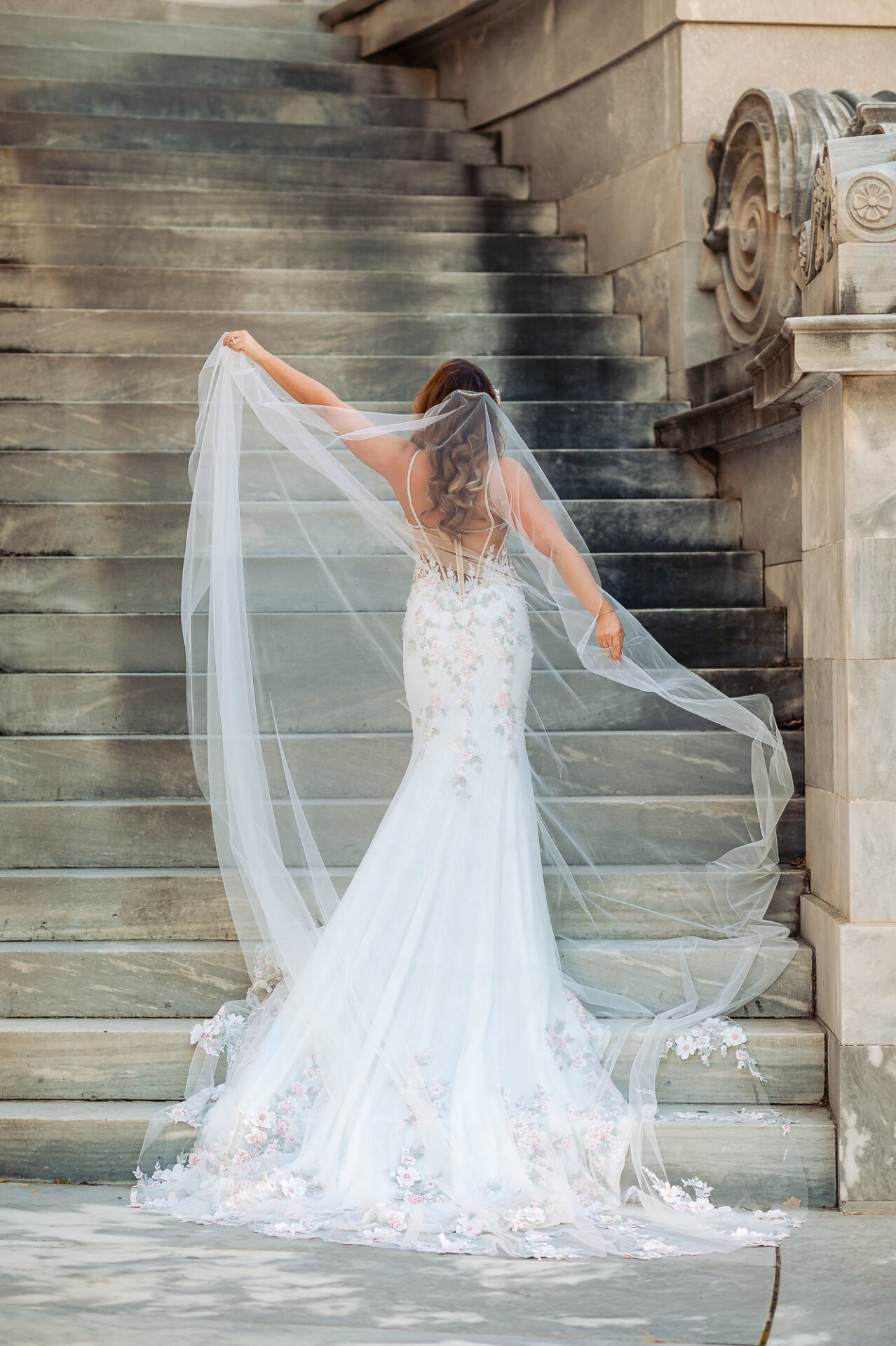 Full rear view of bride walking up stone stairs holding long sheer veil outward
