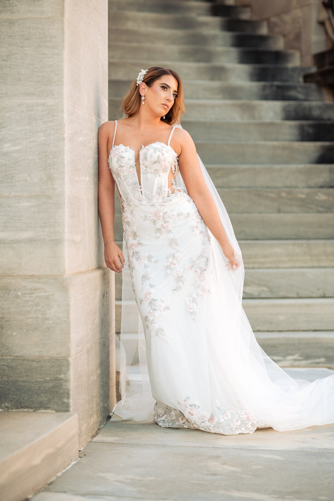 Bride in embroidered fitted gown leaning against marble pillar next to wide stone staircase