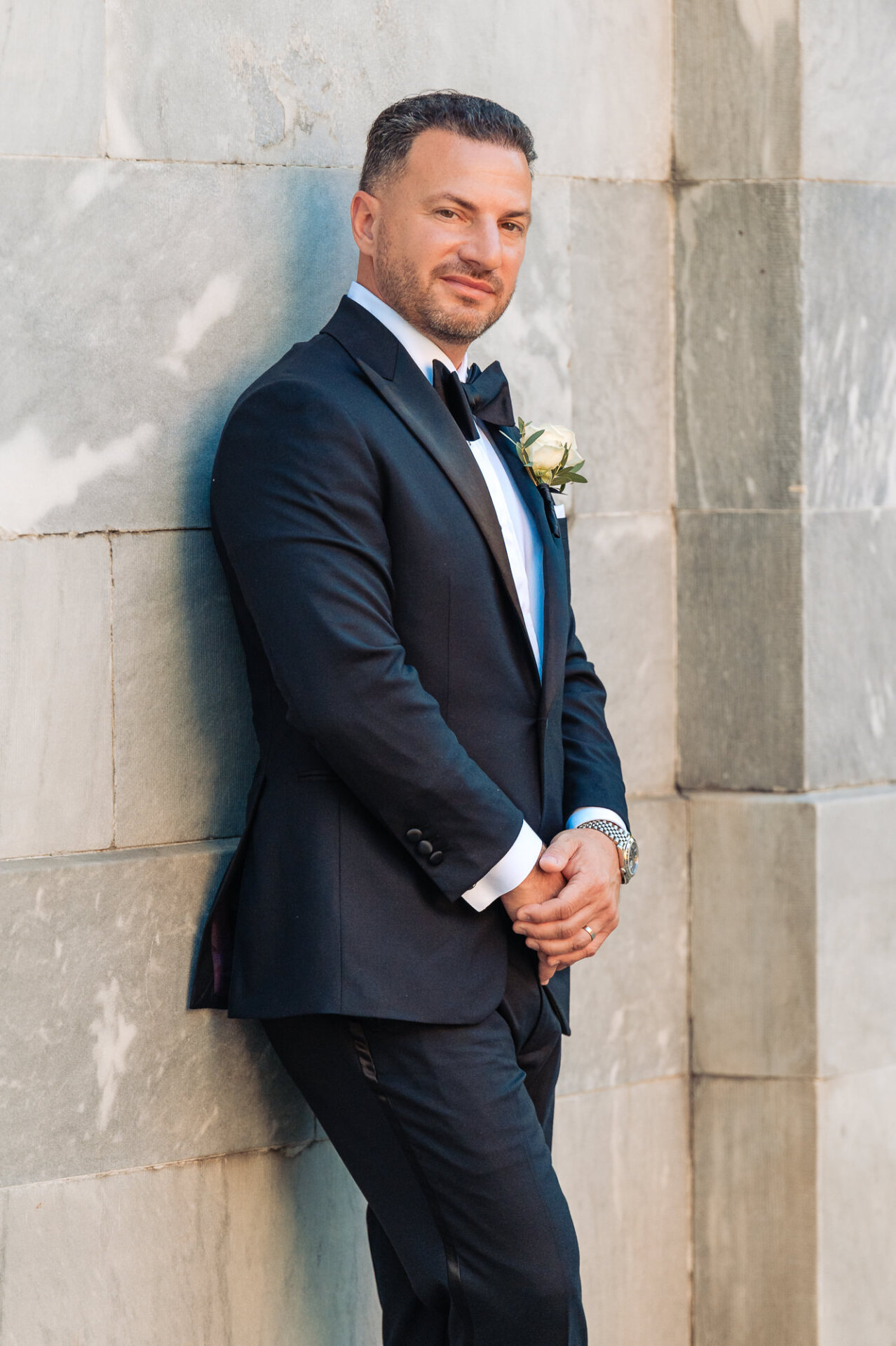 Portrait of groom in black tuxedo leaning against stone wall outdoors looking at camera