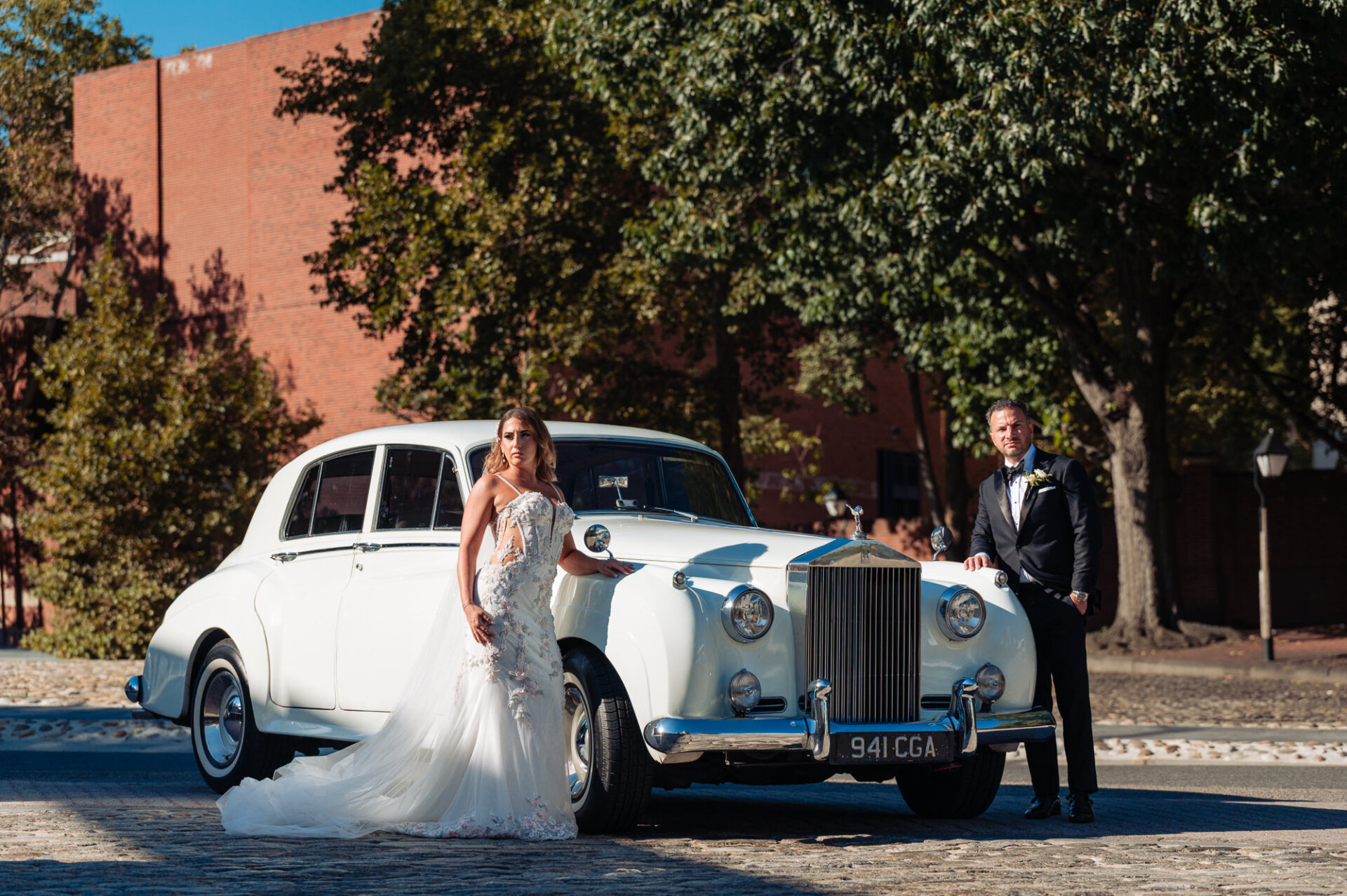 Bride and groom posing outdoors on cobblestone street next to classic white vintage Rolls-Royce wedding car